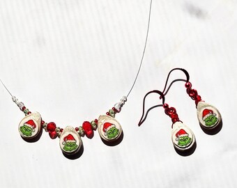 Puede incluir: Un conjunto festivo de collar y pendientes con un diseño del personaje de Grinch. El collar tiene un alambre transparente con colgantes con la cara de Grinch, cuentas rojas y cuentas de cristal transparente. Los pendientes tienen un diseño similar con ganchos rojos.