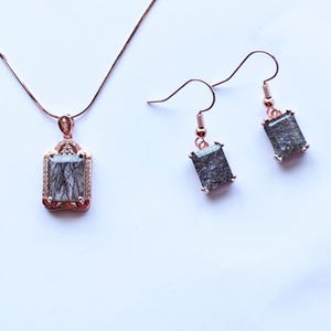 Puede incluir: Un conjunto de collar y pendientes de oro rosa. El colgante y los pendientes presentan piedras rectangulares de color gris oscuro con inclusiones negras, engastadas en un marco decorativo. Las joyas se muestran sobre un fondo blanco.