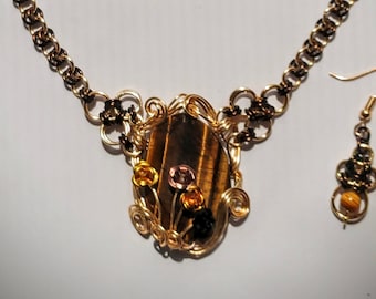 May include: A handcrafted necklace and earring set. The necklace features a gold-toned chain and a pendant with a brown tiger's eye stone, accented with wire-wrapped floral details in various colors. The matching earrings have a similar design.