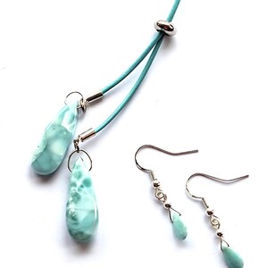 May include: A jewelry set featuring a necklace and earrings. The necklace has a light blue cord with two teardrop-shaped pendants. The earrings have a similar design, with teardrop-shaped stones suspended from silver hooks. The stones have a marbled, light blue and white appearance.