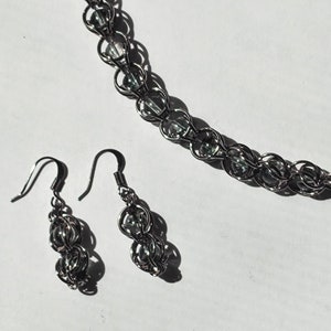 Puede incluir: Un conjunto de joyas plateadas que incluye un collar y pendientes a juego. El collar y los pendientes están hechos de anillos de metal unidos, cada uno con una cuenta redonda y transparente. El conjunto se muestra sobre una superficie blanca.