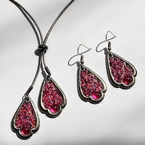 May include: A jewellery set comprising a necklace and earrings. The necklace features two teardrop pendants, and the earrings have a matching design. Each piece has a silver-coloured frame filled with sparkling pink material and a clear pink drop.