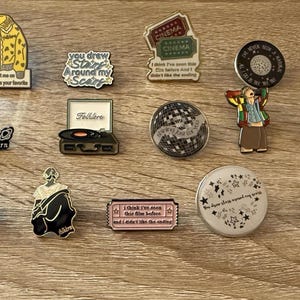 May include: A collection of enamel pins with various designs. The pins include a yellow cardigan, a record player, a disco ball, and pins with text like "love you to the moon and to saturn".