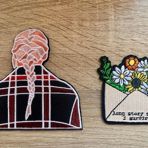 May include: Two embroidered patches. One features a person with long braided hair wearing a red and black plaid shirt. The other patch features a bouquet of flowers in an envelope with the text "long story short, I survived".