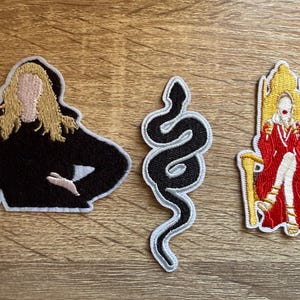 May include: Three embroidered iron-on patches. One features a black silhouette of a woman with blonde hair, another is a black snake, and the third is a woman in a red dress sitting on a gold throne.