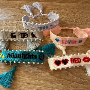 May include: A collection of woven friendship bracelets in various colors, including blue, pink, and white. Each bracelet features embroidered text and decorative tassels. Some bracelets have the words "Midnights", "Lover", "RED", and "1989".