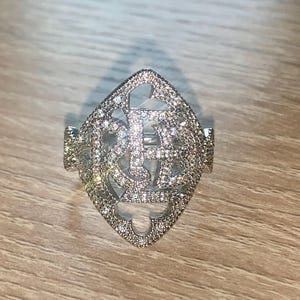 May include: A silver ring with a diamond-encrusted design in the shape of a diamond. The ring is set on a wooden surface.