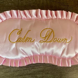 May include: Pink satin sleep mask with gold embroidery that reads "Calm Down".