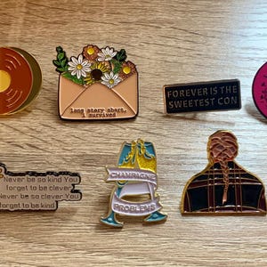 May include: A collection of ten enamel pins featuring various designs, including a record player, a floral arrangement, a calendar, a quote about kindness, a champagne bottle, a person with braids, a Ferris wheel, a bench, and two circular pins with text.
