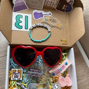 May include: A gift box filled with various items themed around the singer Taylor Swift. The box includes a heart-shaped pair of red sunglasses, a pink bracelet with the word "LOVER" embroidered on it, a small disco ball, a pin with the text "This night is sparkling don't you let it go", a pin with the text "T.S. 1989", a pin with the text "So Long London", a gold necklace with a zodiac symbol, a turquoise beaded bracelet with the text "All Too Well", a sticker with the number "13" on it, and a sticker with a guitar and the text "Folklore" on it.