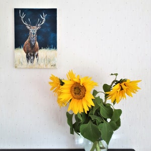 May include: An oil painting of a stag with large antlers against a dark blue background hangs on a white wall. A clear glass vase holds several bright yellow sunflowers with green leaves, sitting on a dark brown surface.