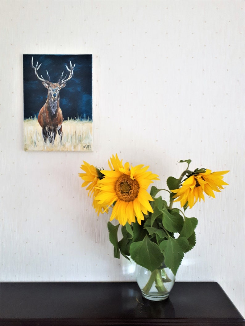 May include: An oil painting of a stag with large antlers against a dark blue background hangs on a white wall. A clear glass vase holds several bright yellow sunflowers with green leaves, sitting on a dark brown surface.