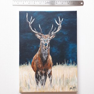 May include: An oil painting on canvas depicts a majestic stag with large antlers, standing in a field of tall grass. The stag has brown fur and a white chest, set against a dark blue background. The painting is approximately 12 inches tall.