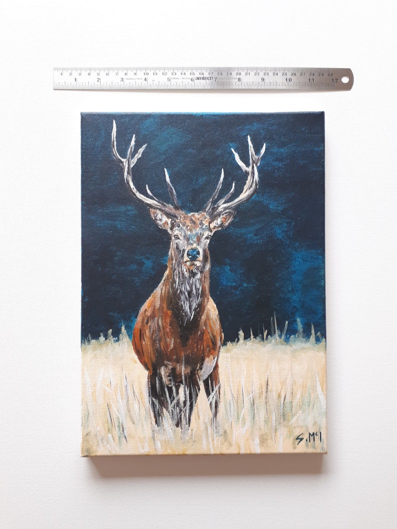 May include: An oil painting on canvas depicts a majestic stag with large antlers, standing in a field of tall grass. The stag has brown fur and a white chest, set against a dark blue background. The painting is approximately 12 inches tall.