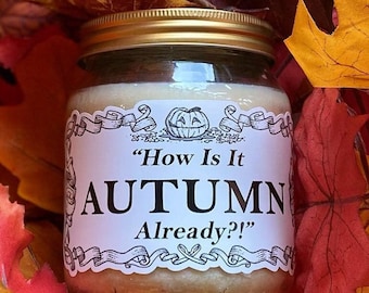 May include: A glass jar candle with a gold lid and a white label. The label reads "How Is It AUTUMN Already?!" with a jack-o'-lantern graphic. The candle is surrounded by colorful autumn leaves.