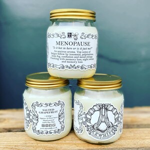 May include: Three glass candle jars with gold lids. The jars are stacked on top of each other. The top jar has a white label with the text "MENOPAUSE" and a humorous description of menopause symptoms. The middle jar has a white label with the text "SALTED GRAPEFRUIT" and information about the candle. The bottom jar has a white label with the text "KEWHIFF" and a graphic of a nose.