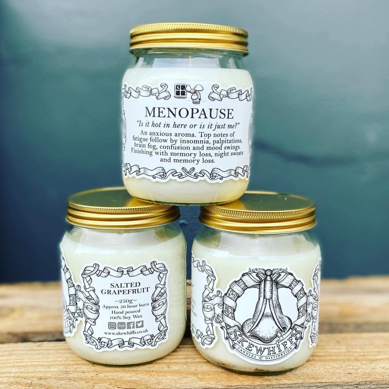 May include: Three glass candle jars with gold lids. The jars are stacked on top of each other. The top jar has a white label with the text "MENOPAUSE" and a humorous description of menopause symptoms. The middle jar has a white label with the text "SALTED GRAPEFRUIT" and information about the candle. The bottom jar has a white label with the text "KEWHIFF" and a graphic of a nose.