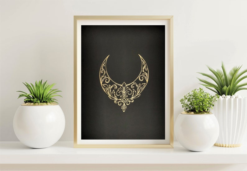 Pu&ograve; includere: Una mezzaluna dorata con intricati disegni floreali su sfondo nero. L'immagine &egrave; incorniciata in una cornice dorata ed esposta su uno scaffale con due piante in vaso bianche.