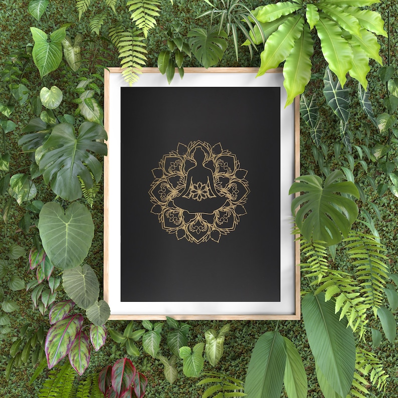 May include: A framed print with a black background and a gold mandala design featuring a person sitting in a meditative pose. The frame is surrounded by lush green foliage.