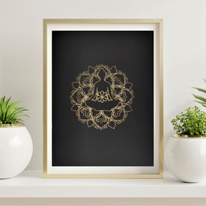 May include: A gold mandala with a silhouette of a person meditating in the center, on a black background. The image is framed in a gold frame and displayed on a white shelf with two potted plants.
