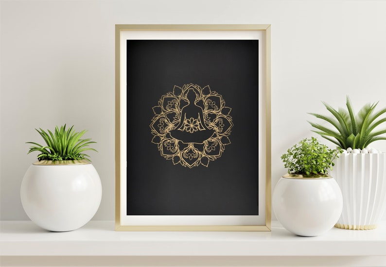 May include: A gold mandala with a silhouette of a person meditating in the center, on a black background. The image is framed in a gold frame and displayed on a white shelf with two potted plants.