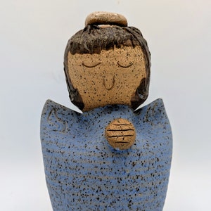 Puede incluir: Figura de ángel de cerámica con vestido azul moteado y halo. El ángel tiene los ojos cerrados y un pequeño adorno redondo y texturizado en el pecho. La figura está hecha a mano y tiene una estética rústica y terrosa.