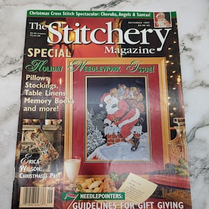 Może przedstawiać: Magazyn The Stitchery, specjalny świąteczny numer poświęcony robótkom ręcznym, prezentuje haft krzyżykowy przedstawiający Świętego Mikołaja schodzącego po kominku. Na okładce znajdują się również skarpetka, poduszki, obrusy i albumy ze wspomnieniami.