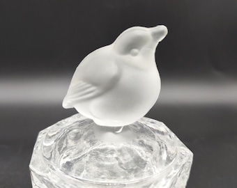May include: A clear glass trinket box featuring a frosted glass bird figurine as the lid handle. The base of the container has a faceted, textured surface, while the bird is smooth and opaque, showing defined wing and beak details.