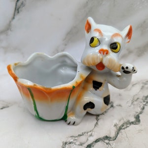 May include: A vintage ceramic planter in the shape of a dog. The dog is white with orange ears and accents, black spots, and yellow eyes. The planter is orange and white with green accents.