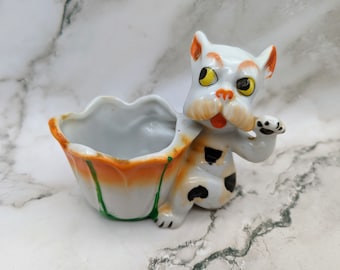 May include: A vintage ceramic planter in the shape of a dog. The dog is white with orange ears and accents, black spots, and yellow eyes. The planter is orange and white with green accents.