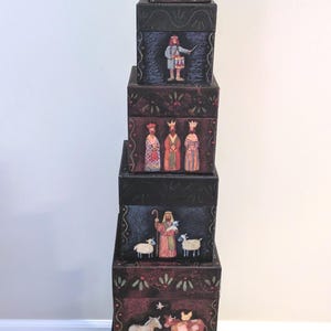 Pode incluir: Uma pilha vertical de seis caixas quadradas pretas, cada uma com ilustrações pintadas à mão. Os desenhos incluem uma estrela dourada, um anjo, um tamborileiro, três figuras com coroas, um pastor com ovelhas, animais de quinta e um presépio.