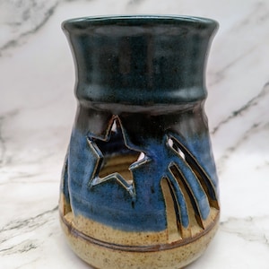 May include: A blue and brown ceramic candle holder with a star cut-out design. The holder has a glazed finish and a rustic look.