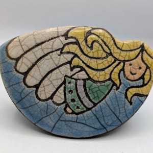 May include: A ceramic angel figurine with a cracked glaze finish. The angel has long yellow hair, a smiling face, and wears a green and purple dress. The wings are white with black outlines, set against a blue background.