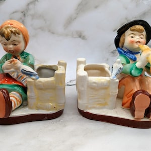 May include: Two vintage ceramic planter figurines. A girl in a red and green dress holds a bird. A boy in a green and blue outfit plays a trumpet. Both figurines are sitting on top of small, rectangular planters.