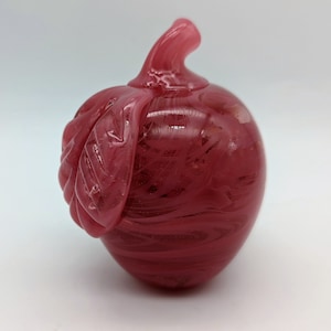 Puede incluir: Una manzana de vidrio rosa con un diseño de espiral y un detalle de hoja. La manzana es un objeto decorativo.