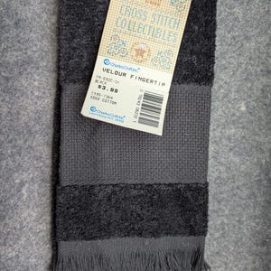 May include: A black cotton velour fingertip towel with a fringed edge and a textured band. A paper tag is attached, featuring the text Cross Stitch Collectibles, Charles Craft Inc., Velour Fingertip, Black, 100% Cotton, and a barcode.