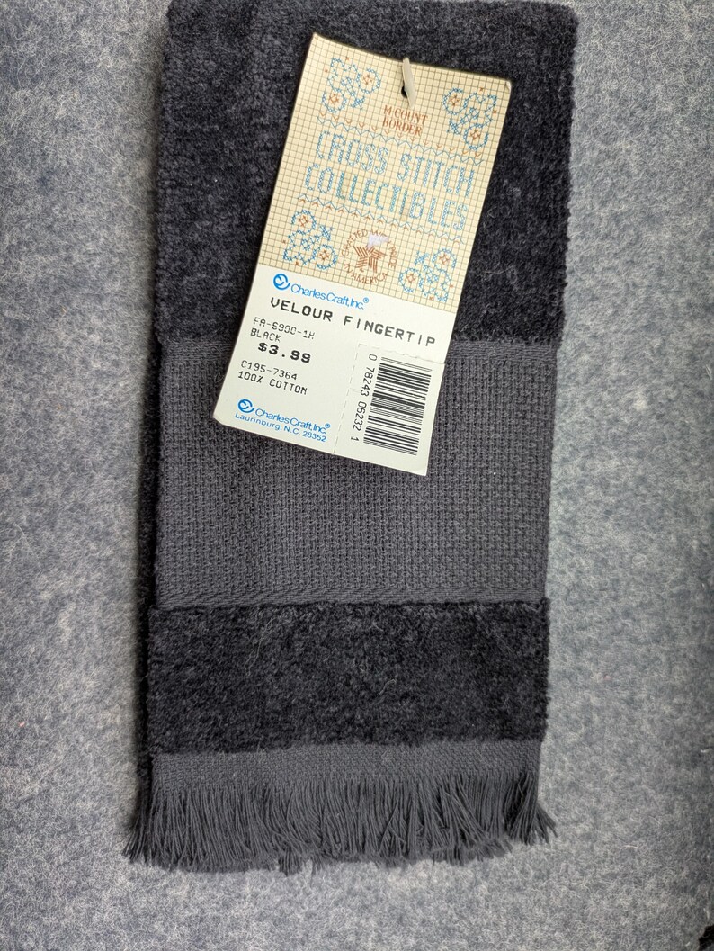 May include: A black cotton velour fingertip towel with a fringed edge and a textured band. A paper tag is attached, featuring the text Cross Stitch Collectibles, Charles Craft Inc., Velour Fingertip, Black, 100% Cotton, and a barcode.