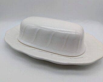 May include: A white ceramic butter dish with a lid, resting on a matching oval-shaped plate. The lid has a ridged design, and the plate has a subtle leaf-like pattern. The dish is designed for serving butter.