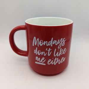Op de afbeelding: Een rode keramische mok met een witte binnenkant en een groot handvat. De mok heeft de witte tekst "Mondays don't like me either". De mok is ontworpen voor warme dranken.