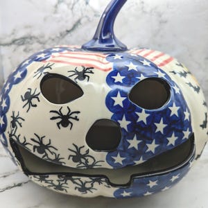 May include: A ceramic jack-o'-lantern featuring a glossy finish with a painted American flag pattern on one side and black spider illustrations on the other. The pumpkin has a dark blue stem, two oval eye cutouts, a nose cutout, and a wide mouth.