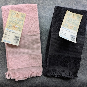 May include: Two velour fingertip towels, one in light pink and one in black, each featuring a textured band and a fringed bottom edge. Both towels have a paper tag attached that reads Cross Stitch Collectibles and Velour Fingertip.