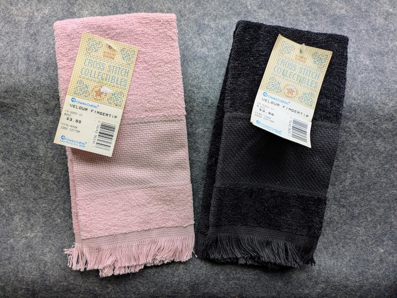May include: Two velour fingertip towels, one in light pink and one in black, each featuring a textured band and a fringed bottom edge. Both towels have a paper tag attached that reads Cross Stitch Collectibles and Velour Fingertip.