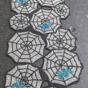 May include: A beaded table runner featuring a repeating pattern of silver spider webs with black outlines. Several webs are accented with small, bright turquoise spiders. The entire piece is constructed from intricate beadwork in a vertical arrangement.