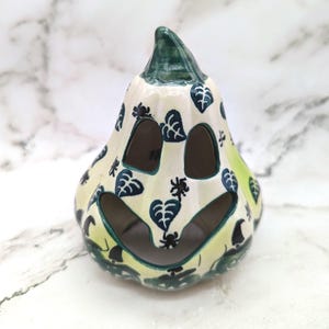 May include: A ceramic pumpkin-shaped lantern with a glossy cream-colored glaze. The surface features hand-painted dark green leaf patterns and small black spider motifs. It has cut-out eyes and a wide mouth, with a green stem at the top.