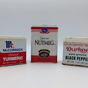 Op de afbeelding: Drie vintage kruidenblikken: McCormick Ground TURMERIC, Kroger Ground NUTMEG en Durkee pure ground BLACK PEPPER. Het kurkumablik is rood en wit, het nootmuskaatblik is beige en rood, en het peperblik is wit en rood.