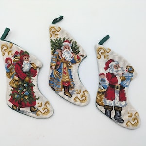 May include: Three needlepoint Christmas stockings featuring detailed Santa Claus figures in various outfits. Each stocking has a cream-colored background with gold scrollwork accents and a dark green fabric hanging loop at the top edge.