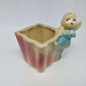 以下が含まれることがあります： 翼のある女の子の形をしたセラミック製のプランターで、ピンクと白のストライプの正方形のプランターを持っています。プランターは釉薬をかけたセラミック製で、薄い青と黄色のカラーリングです。