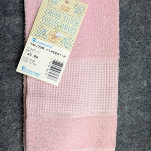 May include: A light pink velour fingertip towel with a textured cross-stitch border section and fringed edges. A paper tag is attached, featuring the text Cross Stitch Collectibles, Charles Craft Inc, Velour Fingertip, Rose, and 100% Cotton.