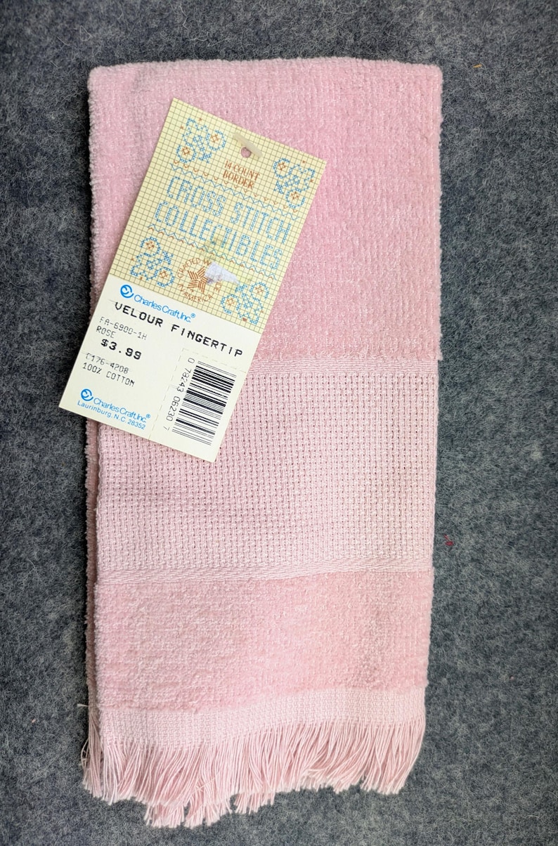 May include: A light pink velour fingertip towel with a textured cross-stitch border section and fringed edges. A paper tag is attached, featuring the text Cross Stitch Collectibles, Charles Craft Inc, Velour Fingertip, Rose, and 100% Cotton.