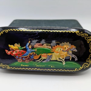 May include: A black lacquered box with a hand-painted scene of a horse-drawn sleigh. The artwork features a vibrant color palette with gold accents and the text "Палех" and "Тройка". The box has a decorative gold border.